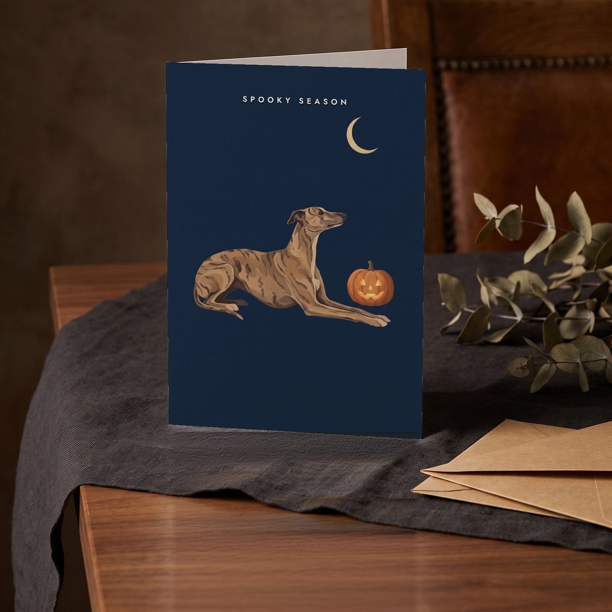 A folded card standing on a charcoal linen cloth with a sprig of dried eucalyptus